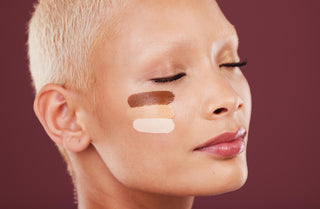 A person with short hair closed their eyes, showing two different shades of foundation on their cheek, against a purple background.