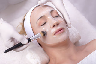 A person is lying down receiving a facial treatment with a mask on their face, being attended to by a professional with gloves and a brush.