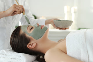A person lying down on a massage table getting a facial treatment, with a green clay mask applied to their face, while a healthcare professional stands beside them holding a bowl.