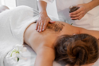 A person lying down on a massage table with coffee scrub applied to their back, being massaged in by another person holding a jar of the scrub.
